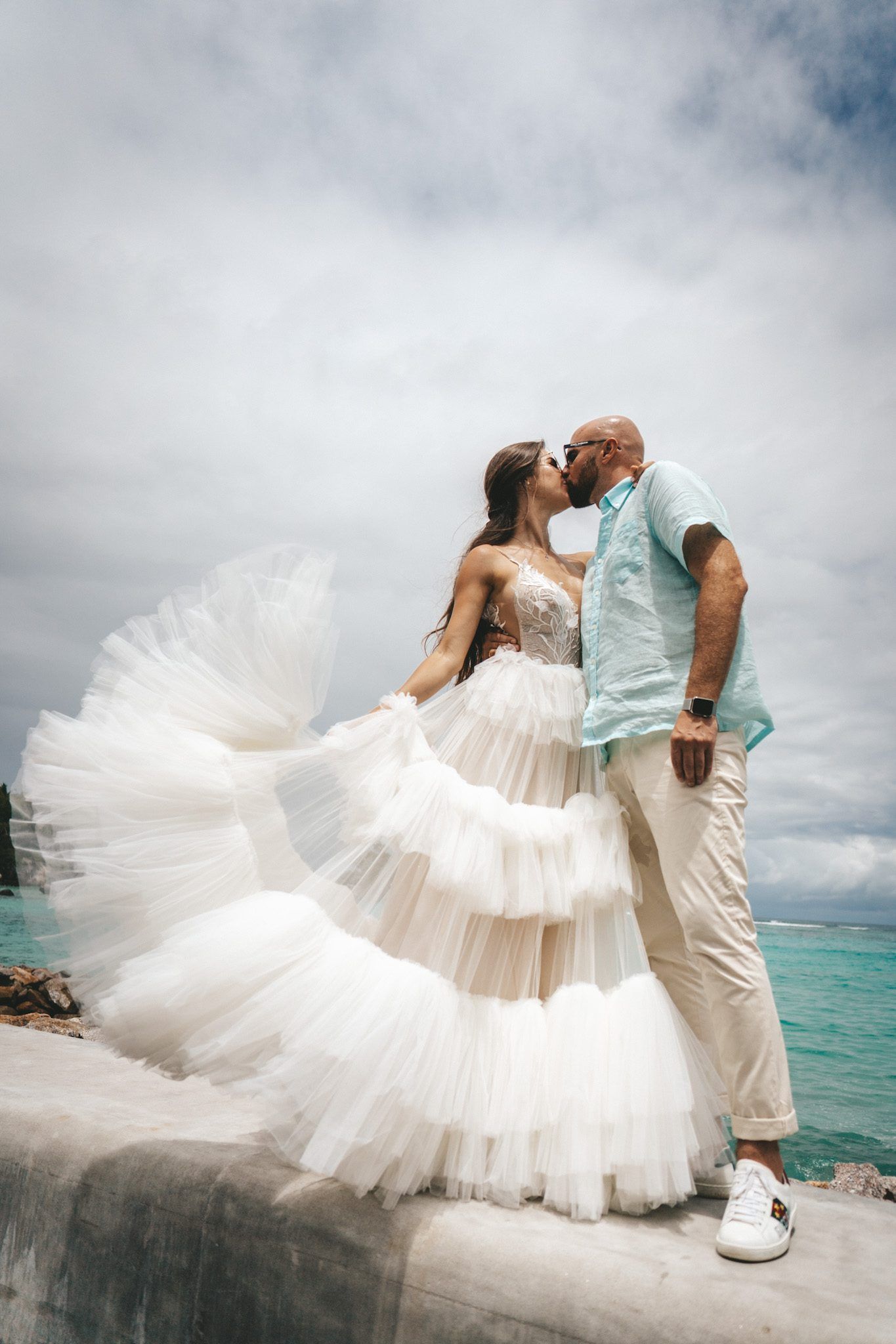Bride in a layered tulle dress kissing the groom on a granite pier above turquoise water