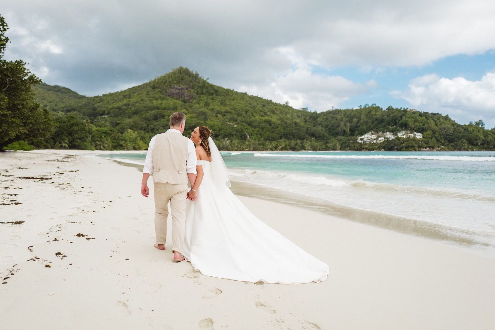 Couple walking barefoot along the shoreline, the bride's train trailing on the sand
