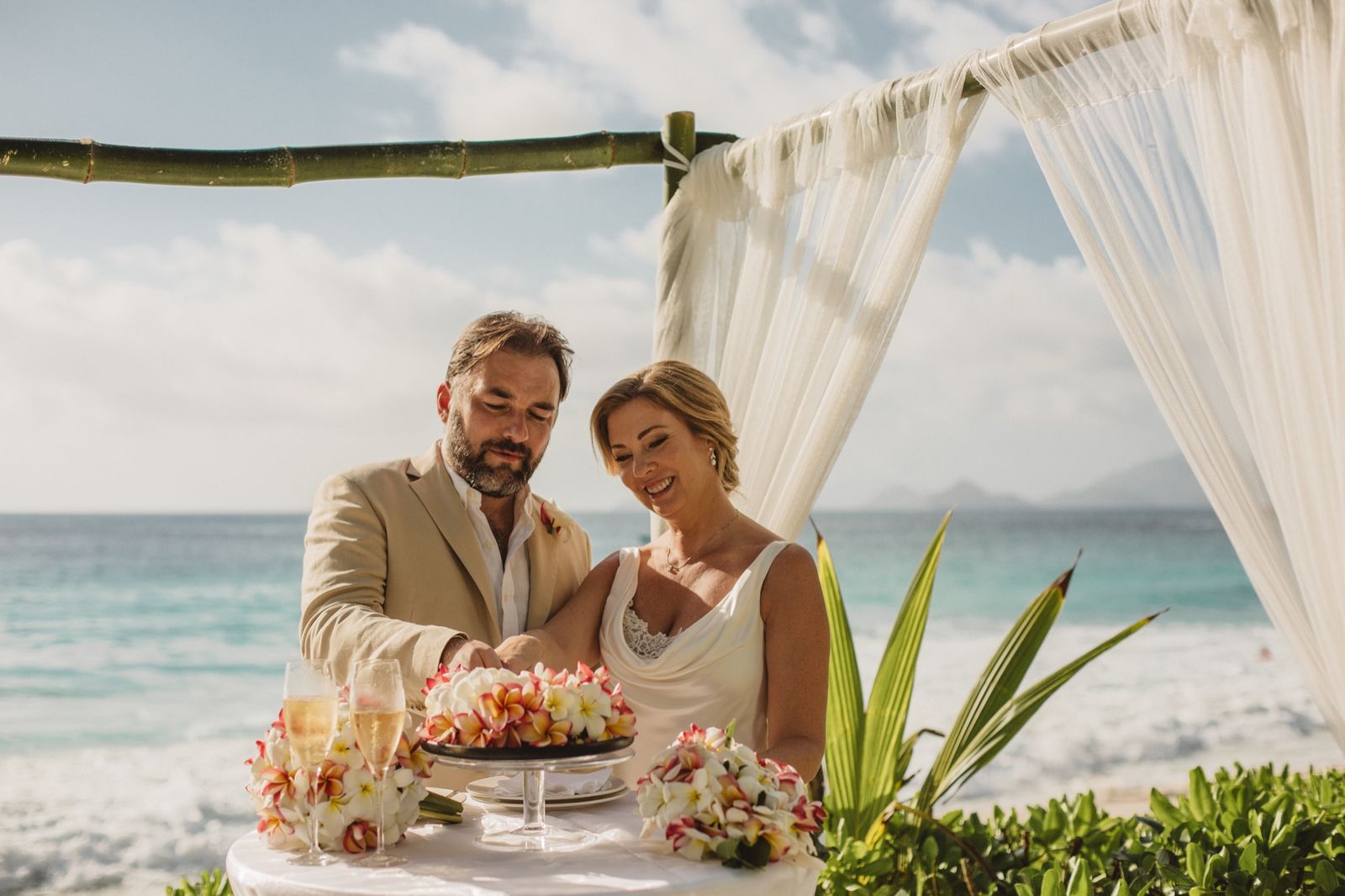 Couple cutting their wedding cake under a bamboo gazebo with white drapes by the sea